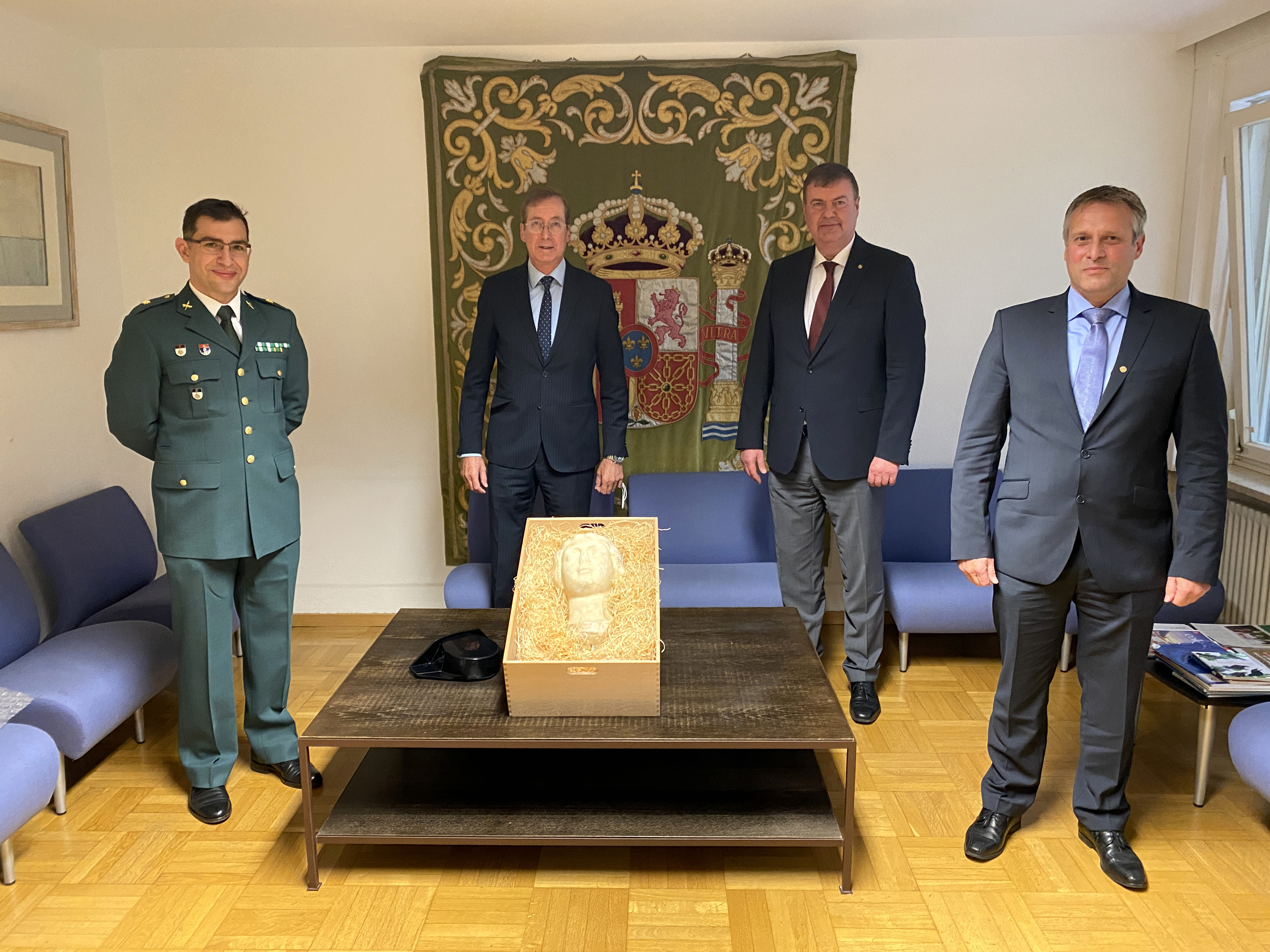A delegation of four men, one of whom is wearing a Greek police uniform, stands around a low wooden table surrounded by a group of sofas, on which a sandstone-colored portrait bust is displayed in a wooden box. A delegation of four men, one of whom is wearing a Greek police uniform, stands around a low wooden table surrounded by a group of sofas, on which a sandstone-colored portrait bust is displayed in a wooden box.